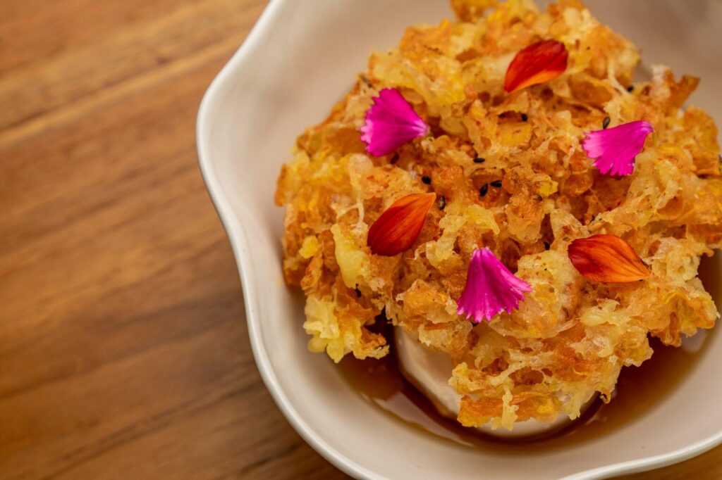 Alt text: A small plate of Marigold Agadeshi, a Japanese tofu dish served in a clear dashi broth with a modern twist. The tofu is delicately fried and topped with crispy fried garlic, green onions, and edible marigold flowers, adding a pop of color to the dish. Served at The Orchid Duck, a Japanese restaurant in Ubud.
